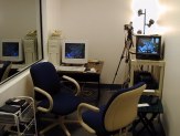 A room with two chairs, a PC, and shelves with a TV and equipment; big mirror on the left